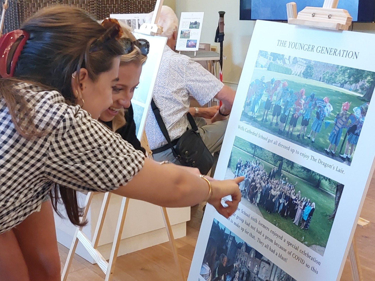 Two young women look at a board with photographs on. They are smiling and pointing.